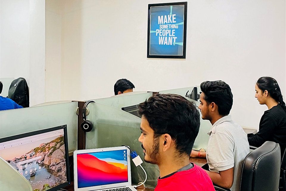 Members at their desks — laptops, monitors, headphones, "Make Something People Want" poster